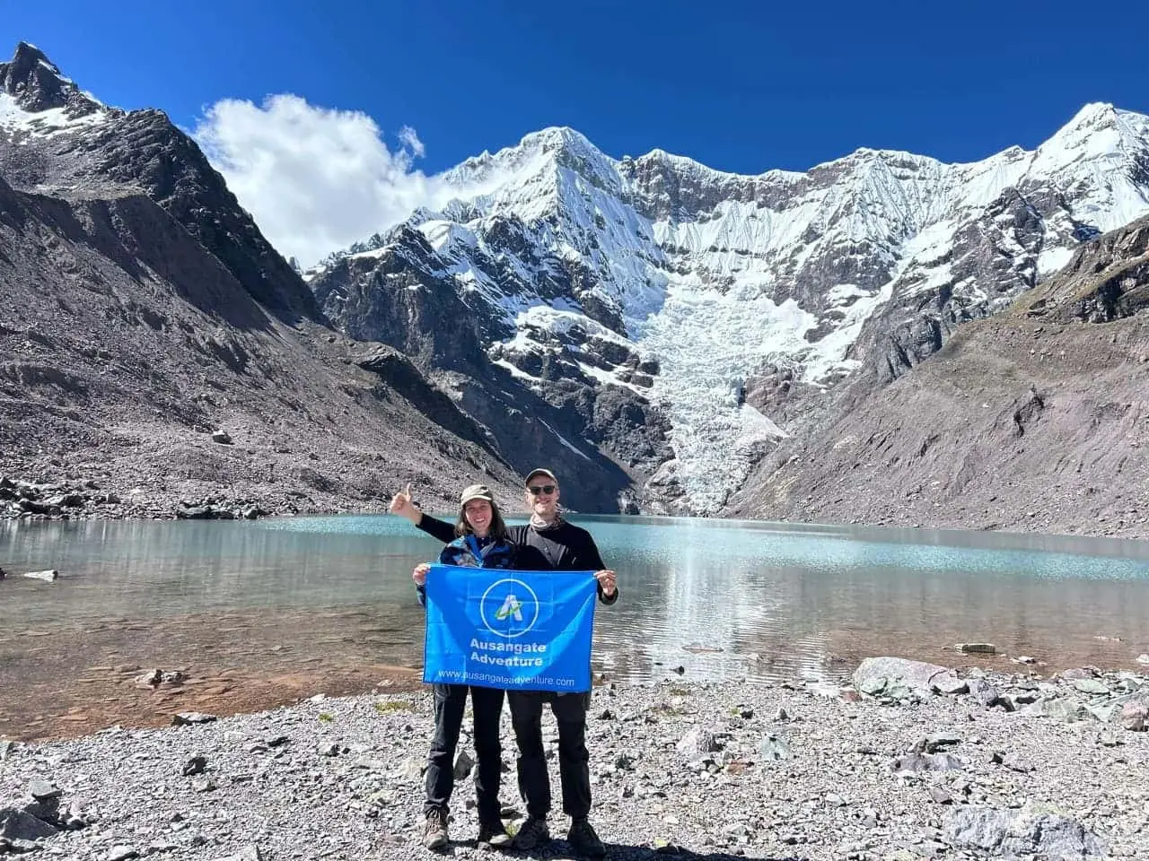 Dos excursionistas sosteniendo una bandera de Ausangate Adventure junto a una laguna glaciar turquesa en el nevado Ausangate, Perú.