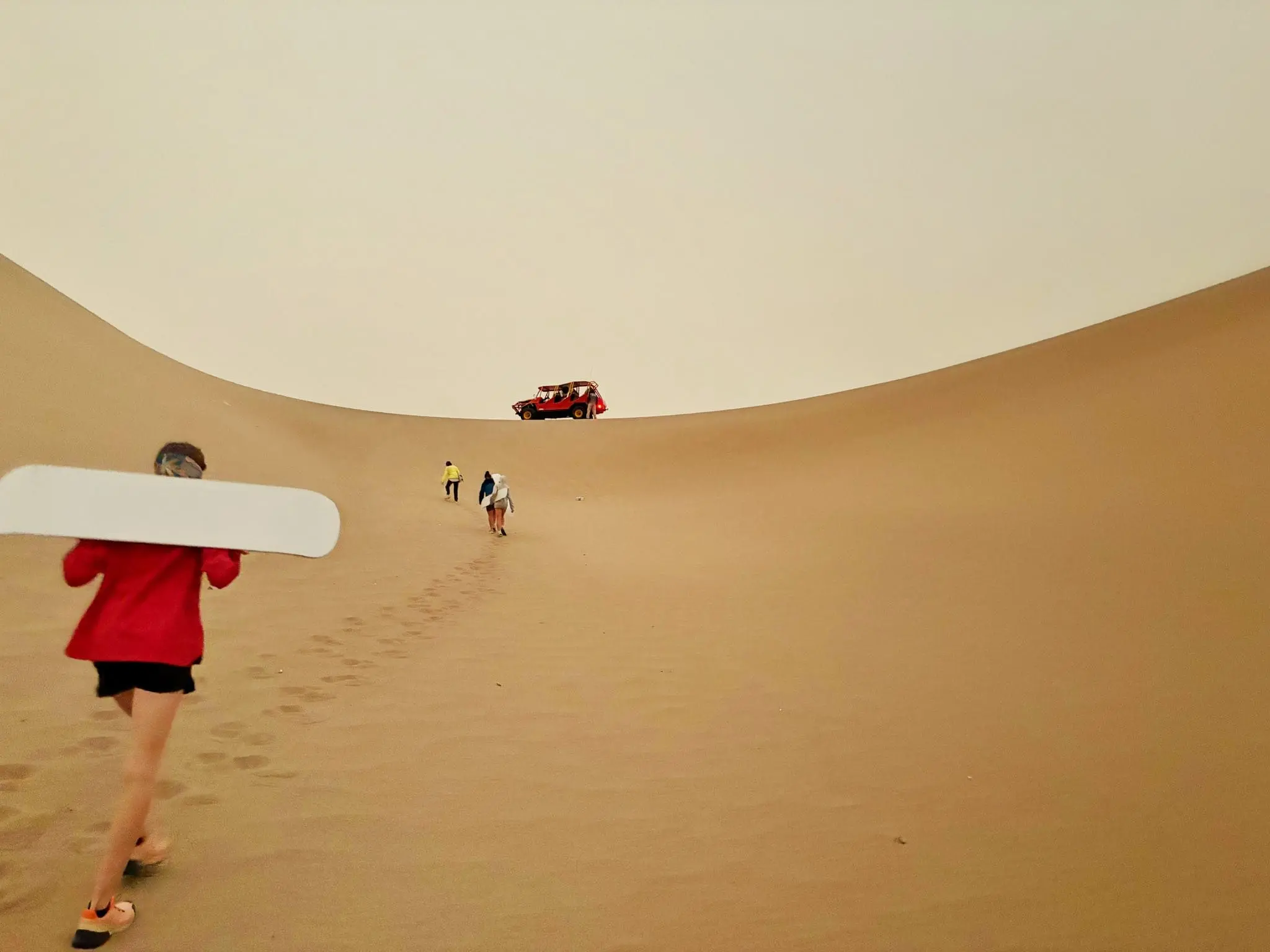 Hiker with a sandboard walking up a desert sand dune toward a red buggy in Huacachina, Peru