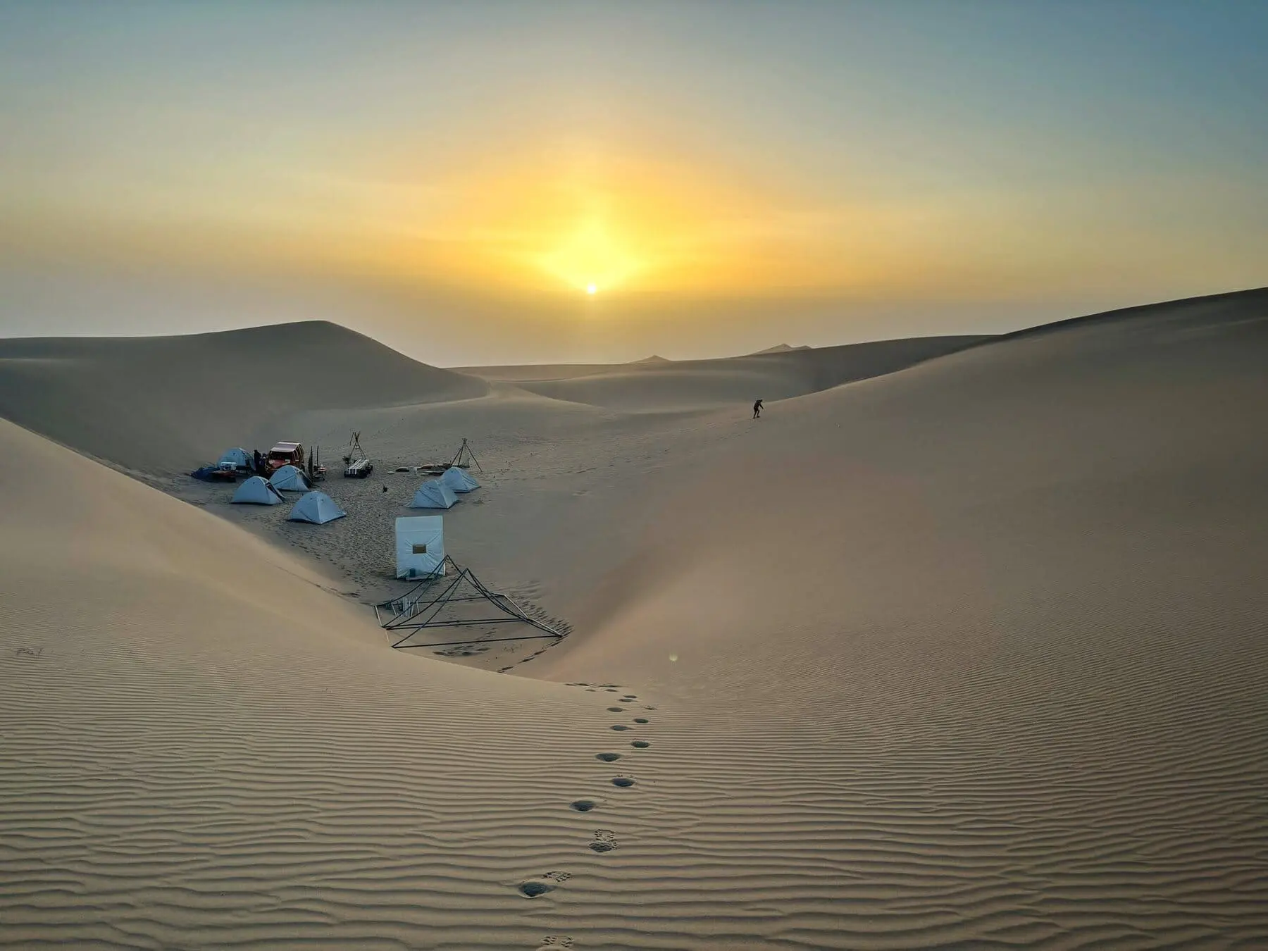 Footprints in the sand leading to a desert camp setup during a golden sunset in Huacachina, Peru.