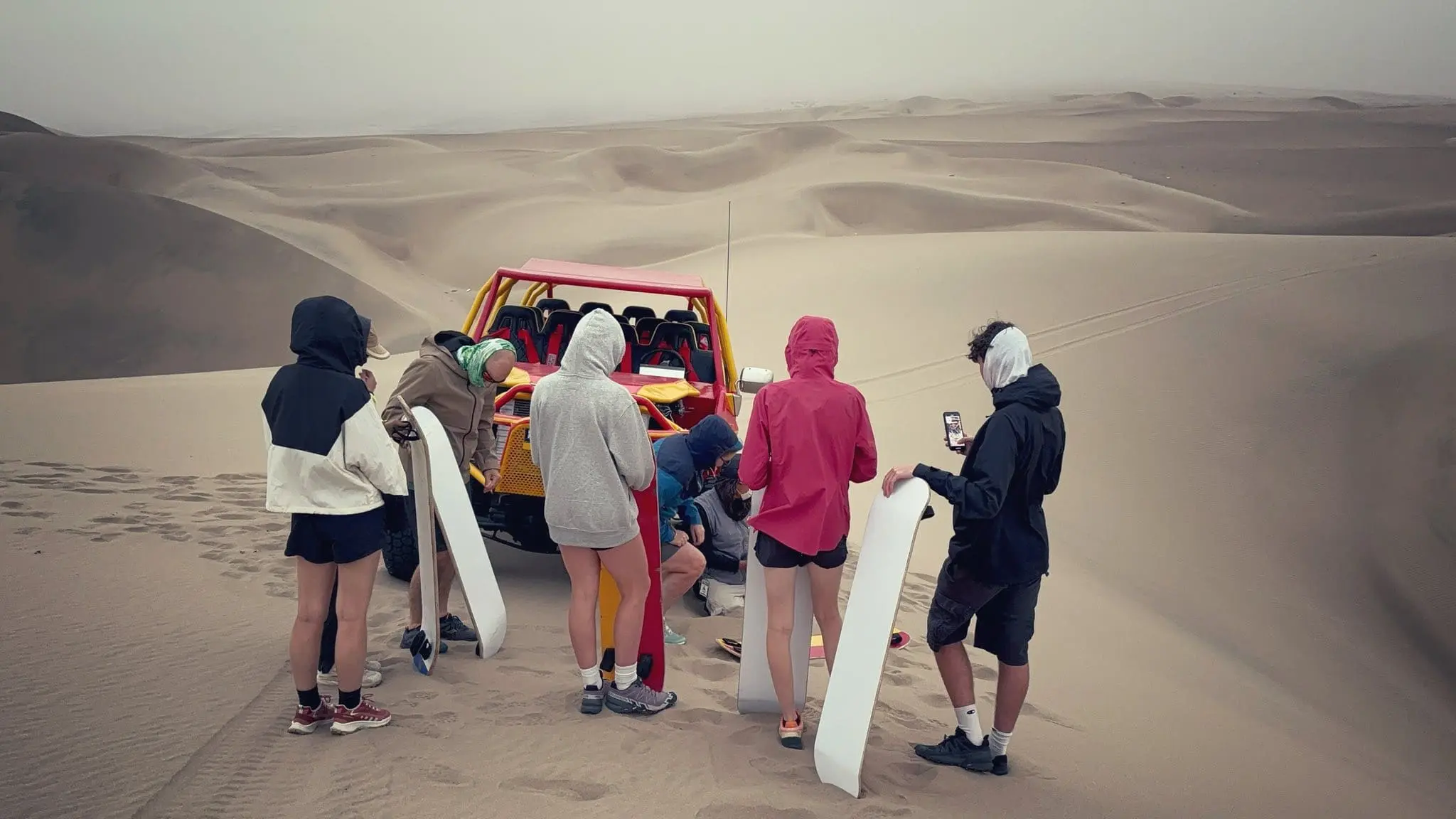 A group of travelers with sandboards standing next to a red dune buggy in the Huacachina desert, Peru