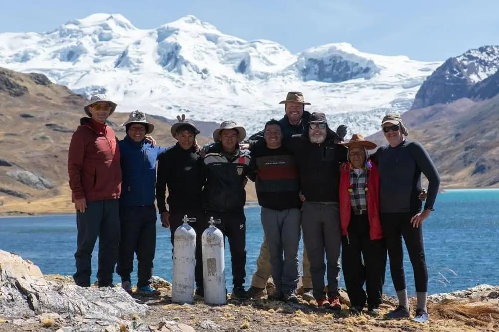 Group of trekkers and guides posing beside Sibinacocha Lake with the snowcapped Ausangate Mountain in the Peruvian Andes.